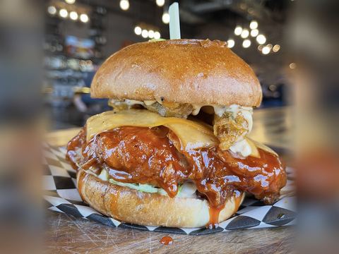 The Nashty Hot Chicken: a crispy fried chicken thigh in glossy red hot sauce with melted cheddar, fried pickles, creamy sauce and lettuce on a toasted brioche bun with a skewer, on checkered paper on a wooden table